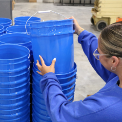 Employee inspecting blue food-grade buckets in warehouse, selecting quality containers from stacked inventory
