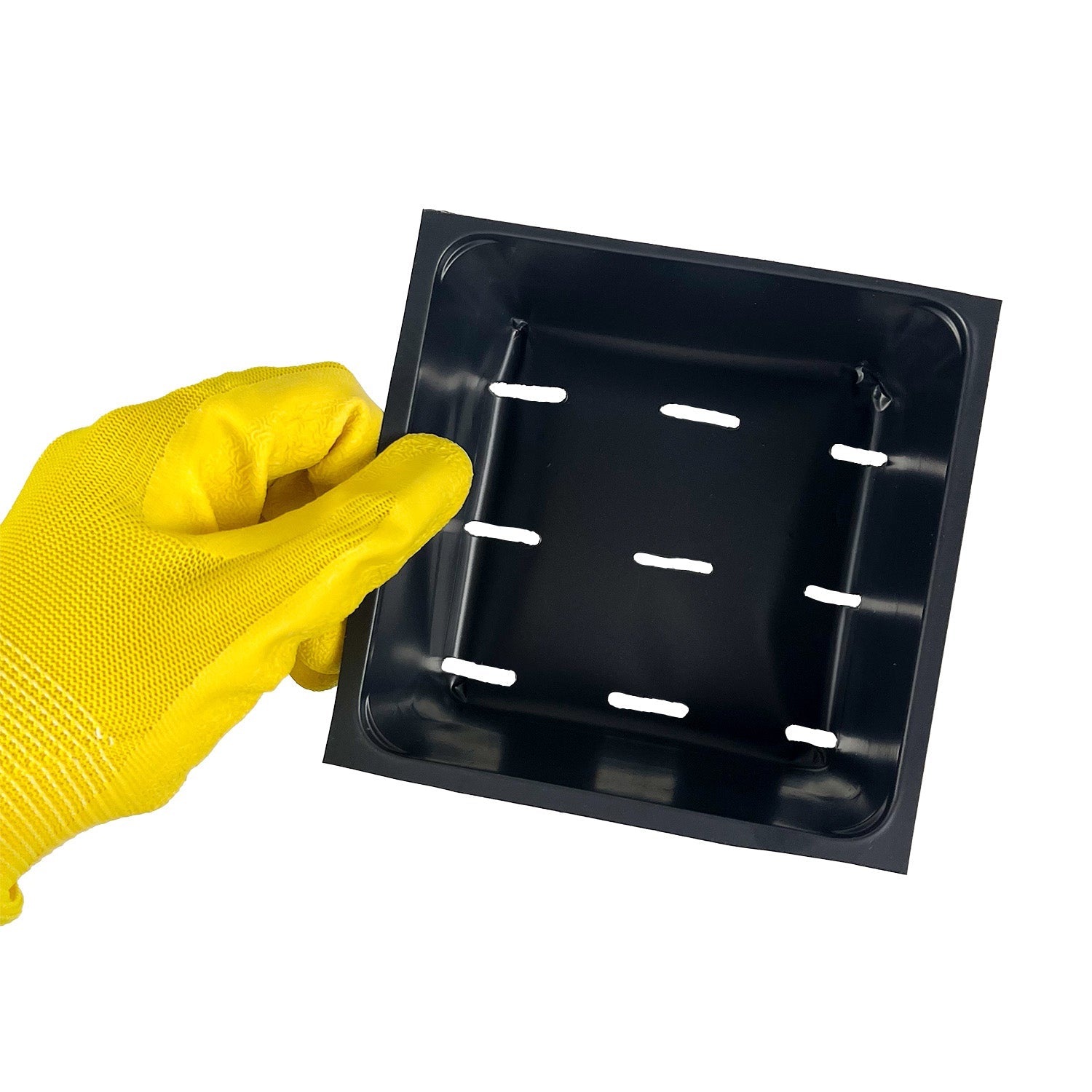 A hand in a yellow glove holds a black square tray with six rectangular ventilation holes evenly spaced in the center.