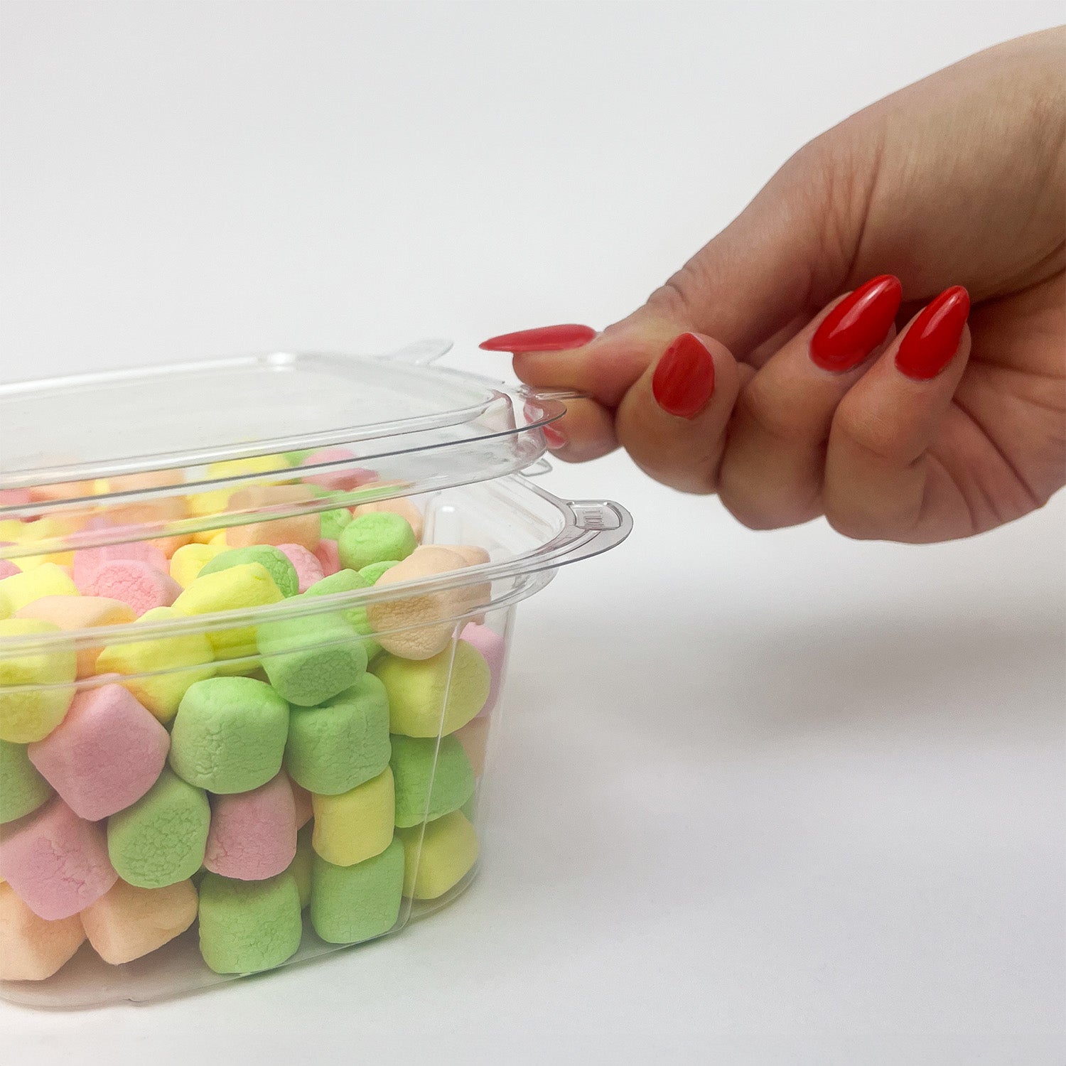 A hand with bright red nails opens a 16 oz. Tamper-Evident Container - Crystal Seal, filled with pastel-colored marshmallows, against a plain white background.