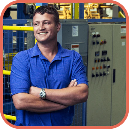 A man in a blue polo shirt stands with arms crossed, smiling, in front of industrial equipment and control panels inside a factory or manufacturing facility.