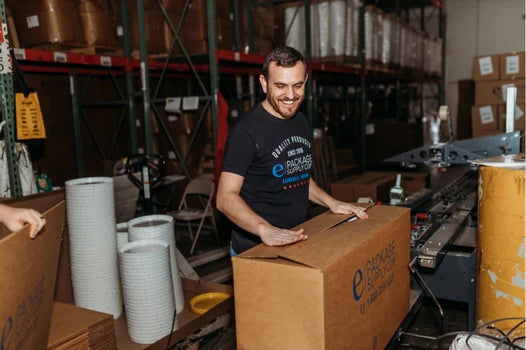 A man smiles while packing a large cardboard box in a warehouse, surrounded by shelves of supplies and other packaging materials.