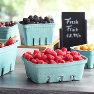 Small blue cartons filled with fresh raspberries, blackberries, grapes, and other fruit sit on a counter with a chalkboard sign in the background that reads Fresh Fruit and a price.