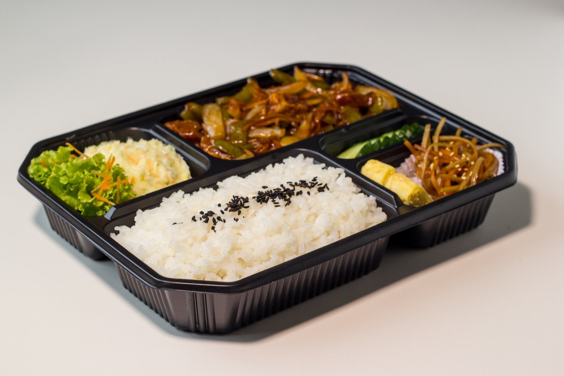 A black bento box with white rice topped with black sesame seeds, stir-fried vegetables, a leafy green salad, pickled vegetables, and a rolled omelette on a light background.