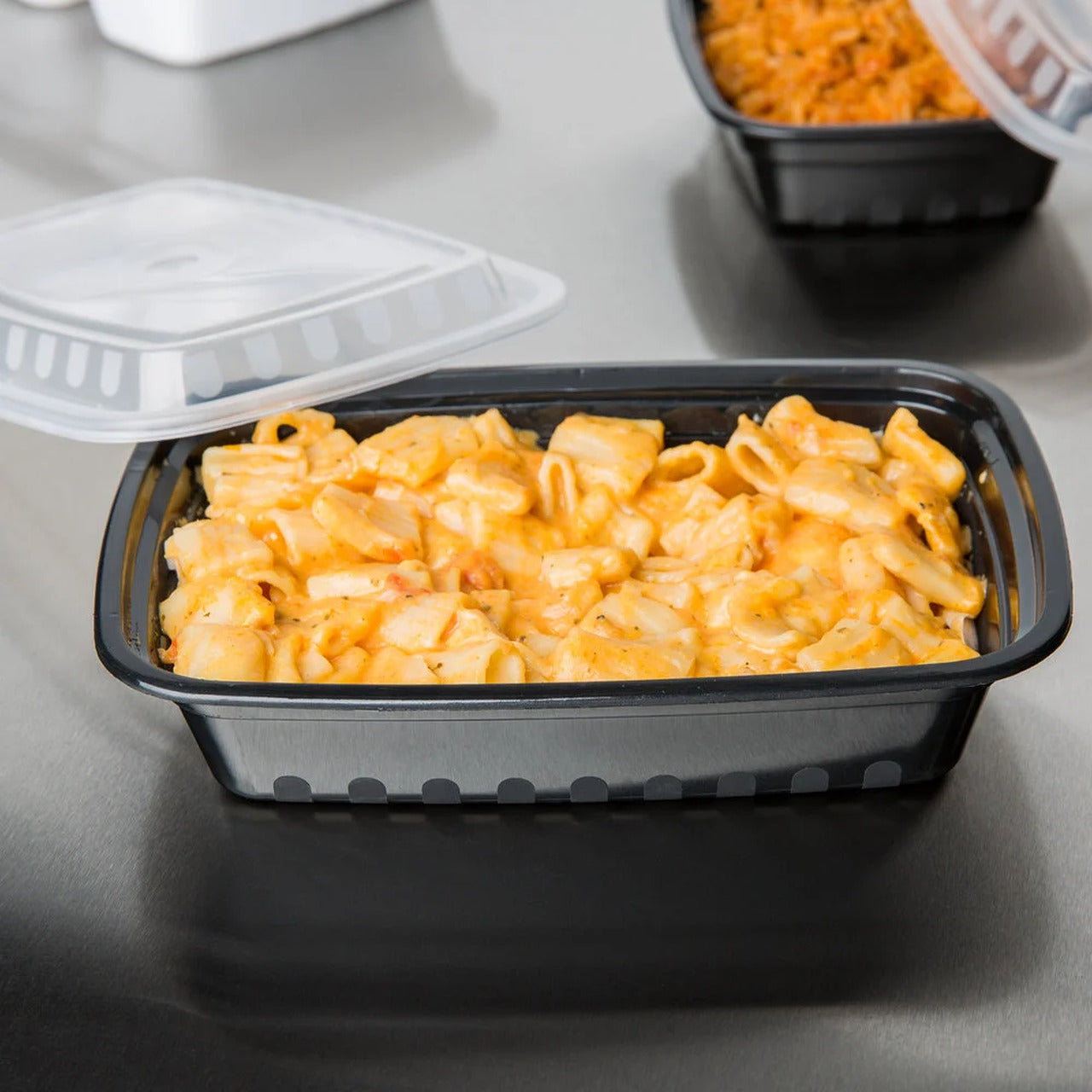 A black plastic food container filled with creamy macaroni and cheese sits on a stainless steel surface, with its clear plastic lid partially open.