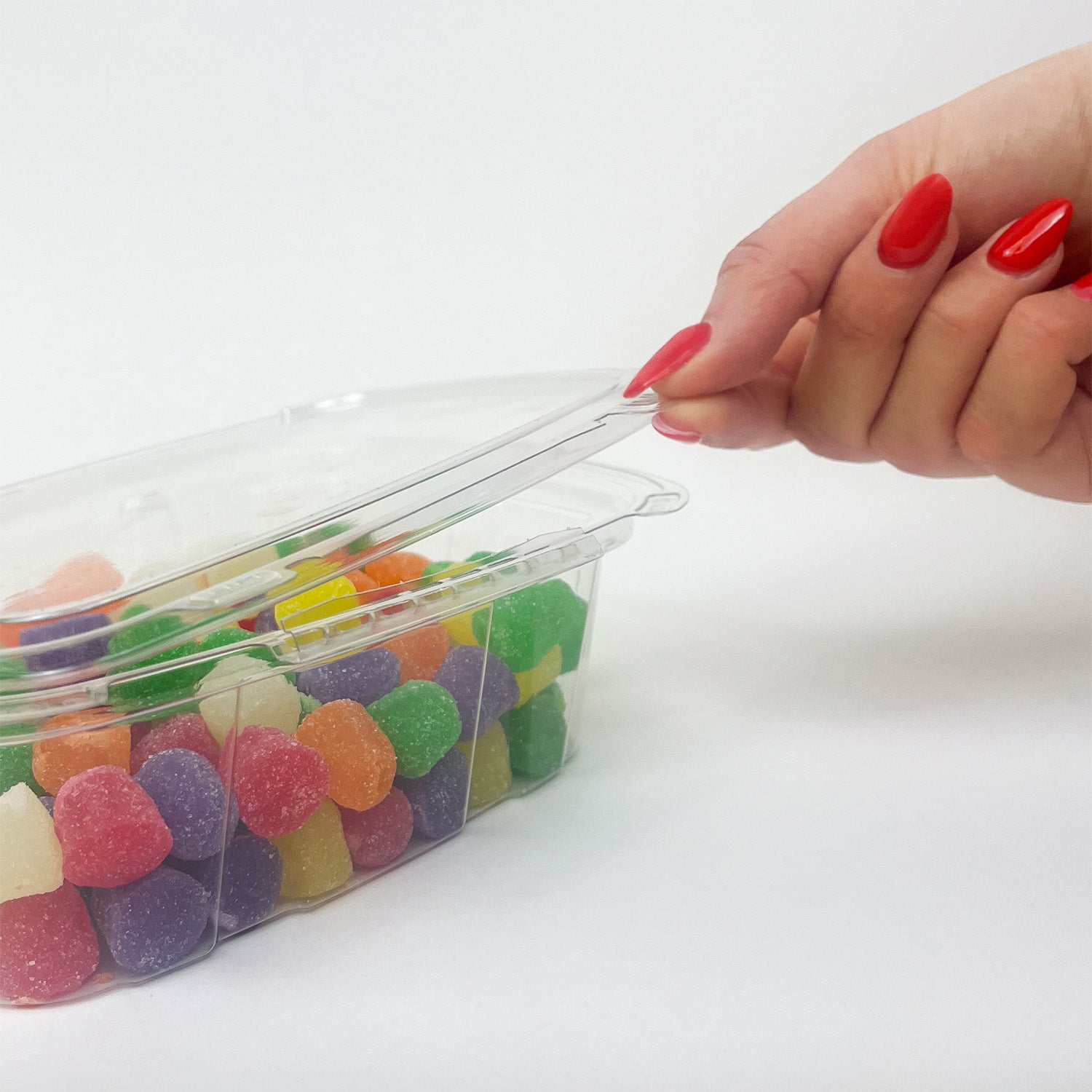 A hand with red painted nails lifts the lid of a Crystal Seal 24 oz. Tamper-Evident Container filled with colorful gumdrops, shown against a plain white background.