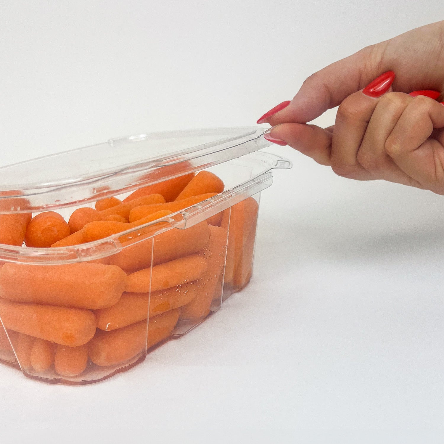 A hand with red-painted nails opens a Crystal Seal 32 oz. Tamper-Evident Container (case of 164), filled with baby carrots, against a plain white background.