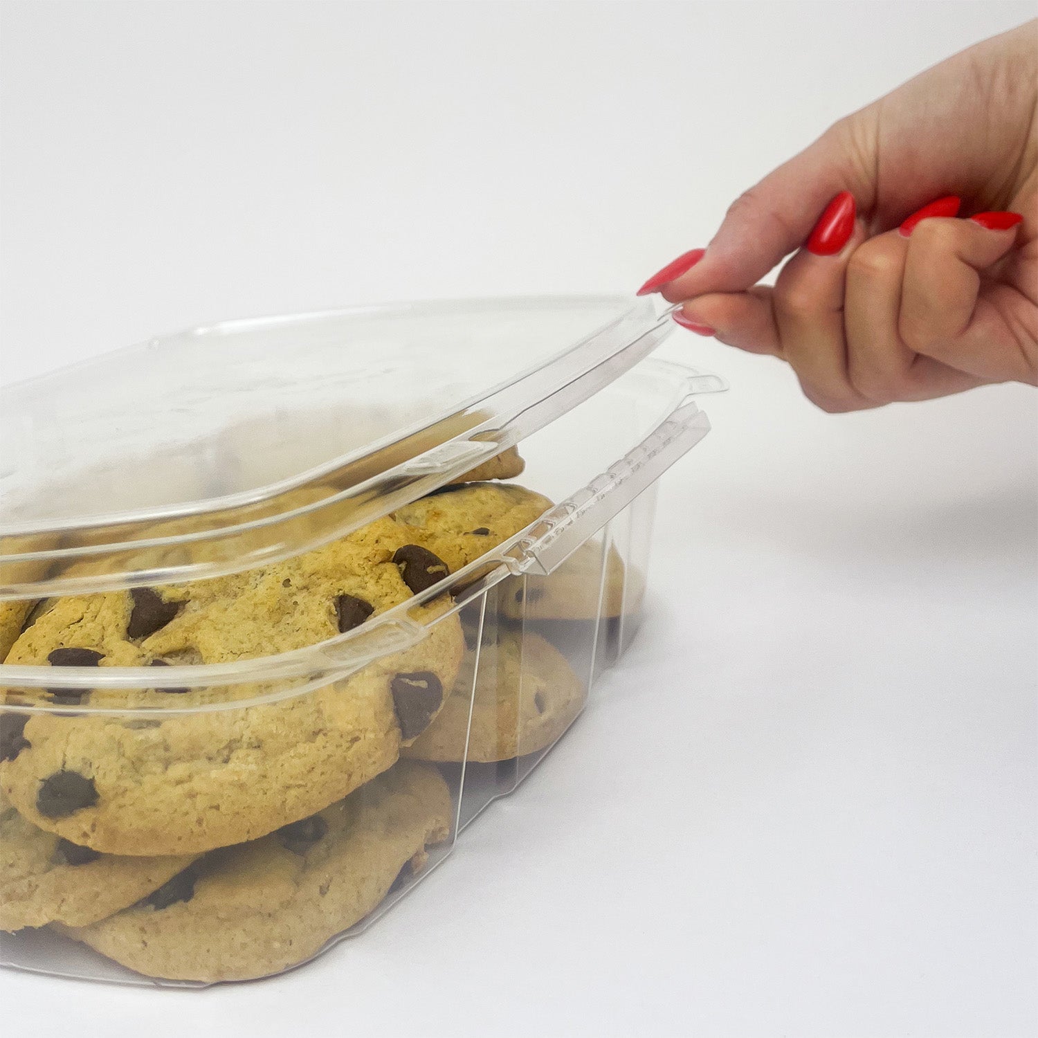 A hand with red painted nails opens a 48 oz. Tamper-Evident Container - Crystal Seal, case of 140, filled with chocolate chip cookies against a plain white background.