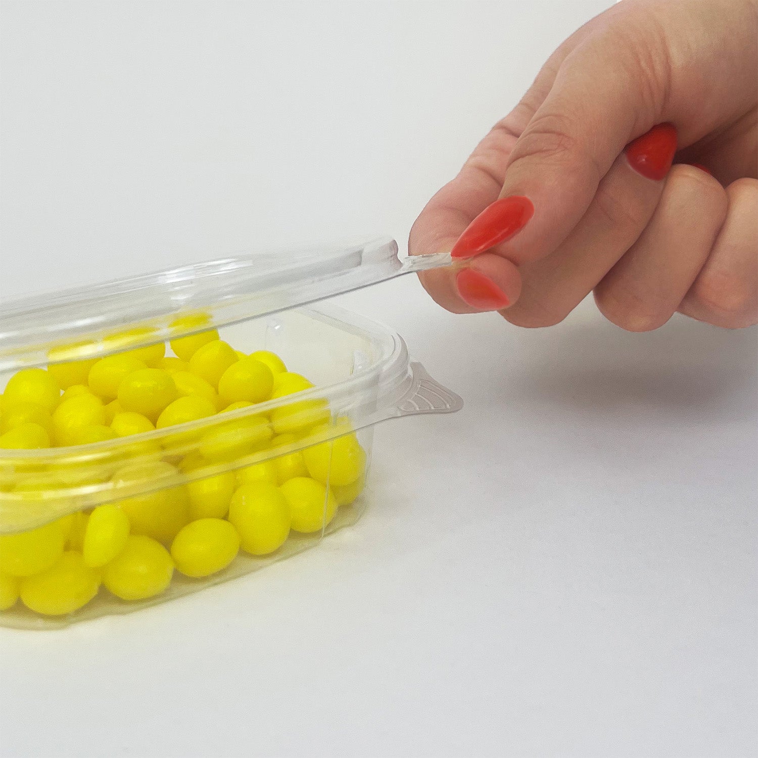 A hand with red-painted nails opens a 4 oz. Crystal Seal Hinged Deli Container (case of 400), filled with small, round yellow candies, on a white surface.
