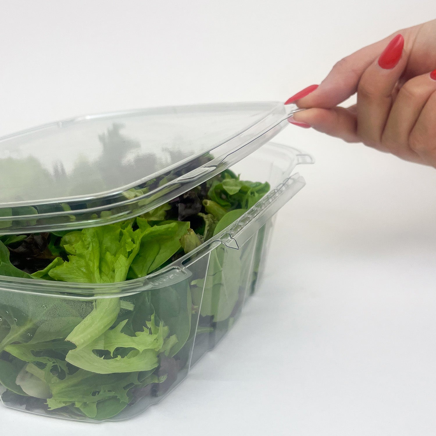 A hand with red nail polish lifts the lid of a 64 oz. Tamper-Evident Crystal Seal container, case of 140, shown filled with mixed salad greens against a plain white background.