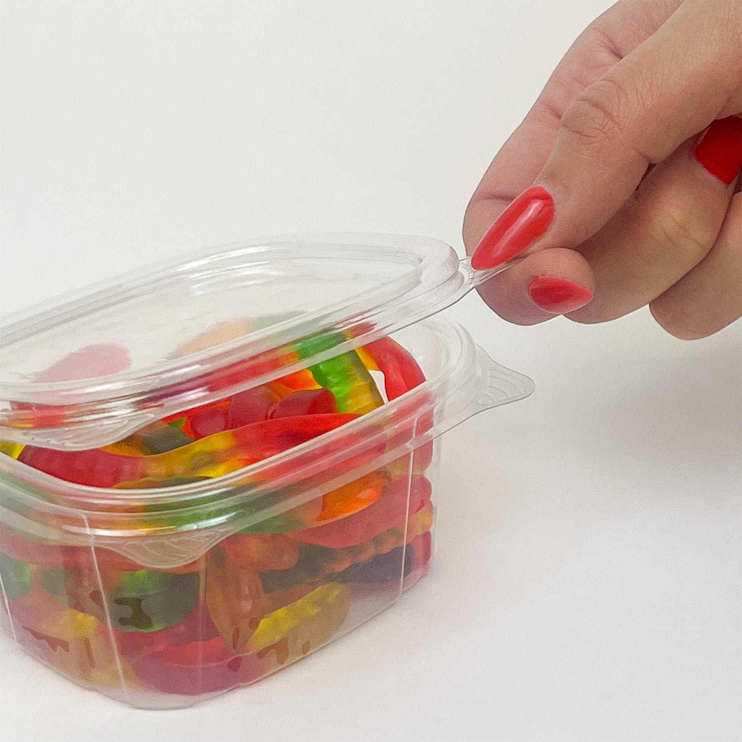 A hand with red-painted nails opens a 6 oz. Crystal Seal Hinged Deli Container, revealing colorful gummy bear candies inside, set against a plain white background.